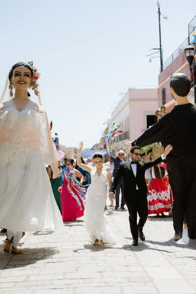 Boda en Oaxaca: Fotografía de una pareja de novios caminando entre dos figuras de ellos hechas de cartón tamaño grande.