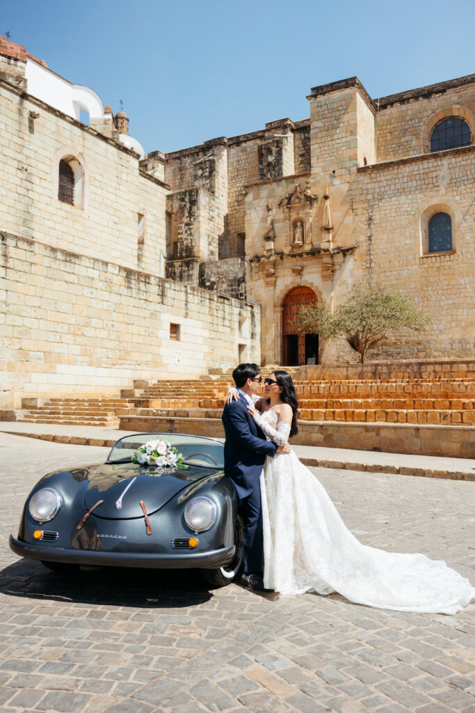 Boda en Oaxaca: Fotografía de una pareja de novios de pie a lado de un automóvil clásico color negro.