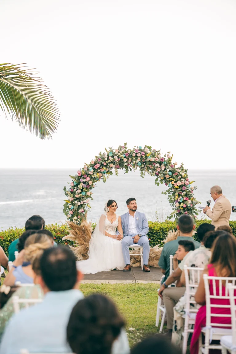 Funciones de un wedding planner: Fotografía de una pareja sentados al aire libre bajo un arco de flores.